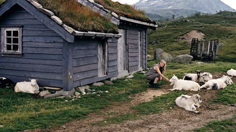 En dame sitter foran en hytte på fjellet og klapper på en geit.