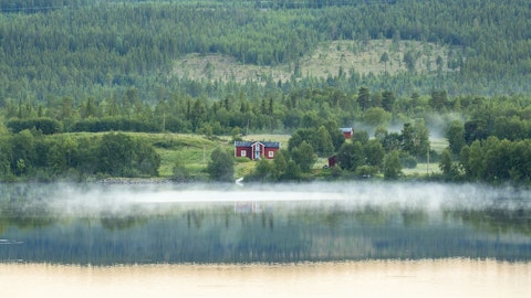 Utsikt over vann, på andre siden er det et rødt hus med love og skog