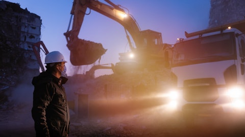 En mann med maske står foran en gravemaskin og en lastebil i halvmørket.